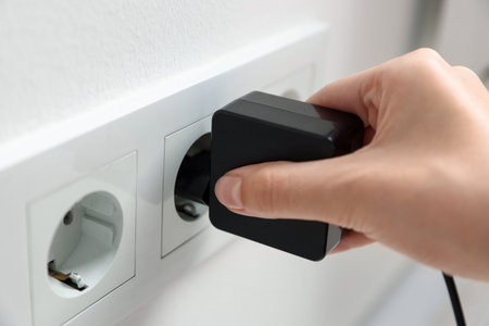 Woman plugging USB adapter with cable into electric socket on white wall, closeup