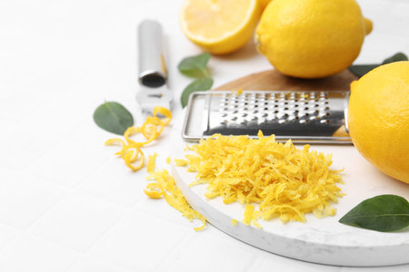 Lemon zest, fresh fruits, tools and green leaves on white tiled table, closeup