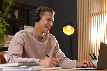 Man in headphones taking notes while passing online test via laptop at table indoors