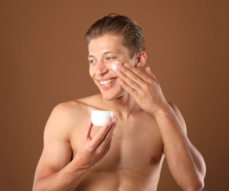 Young man applying cream onto his face on brown backgroundの写真素材