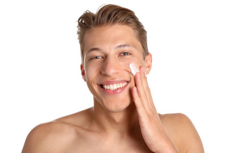 Handsome young man applying cream onto his face on white backgroundの写真素材