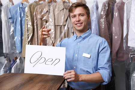 Dry-cleaning service. Happy worker holding Open sign at near rack with clothes indoors