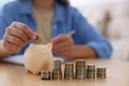 Woman putting coin into piggy bank at wooden table indoors, closeup