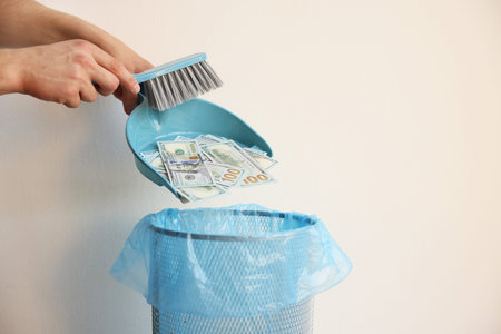 Man throwing dollar banknotes from dustpan into trash bin near white wall, closeup. Space for textの写真素材