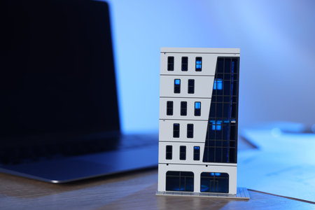 Real estate. Apartment building model and laptop on wooden table in color light, closeup