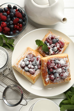 Tasty puff pastries with berries, powdered sugar and mint on white wooden table, flat lay