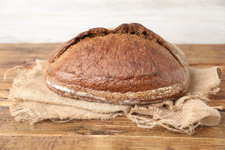 Loaf of fresh bread and burlap cloth on wooden table against light background, closeup