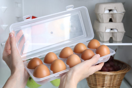 Woman holding container with raw chicken eggs near refrigerator, closeup