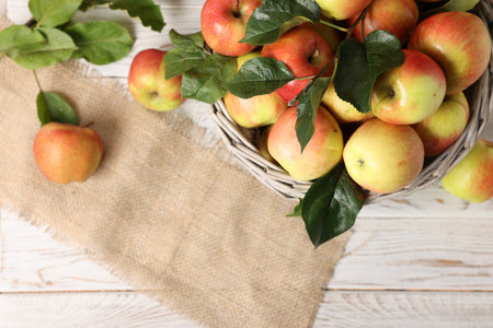 Fresh ripe apples with green leaves in wicker basket on white wooden table, top view. Space for textの写真素材