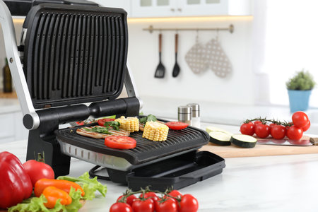 Electric grill with tasty meat and vegetables on white marble table in kitchen