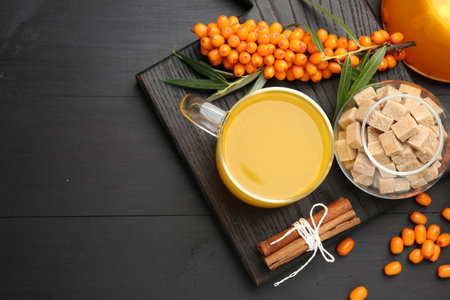 Sea buckthorn tea in glass cup, fresh berries, brown sugar and cinnamon on black wooden table, flat...