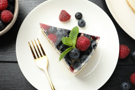 Piece of delicious cheesecake with berries and mint on black wooden table, flat lay