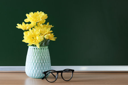 Happy Teacher's Day. Vase with flowers and glasses on wooden table near chalkboard, space for textの写真素材