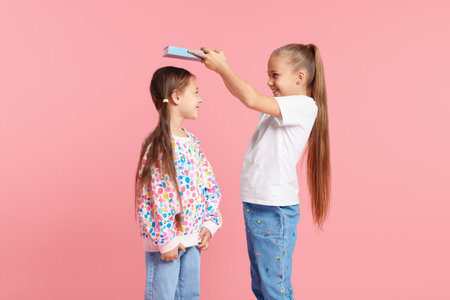 Little girl with book measuring her friend's height on pink backgroundの写真素材