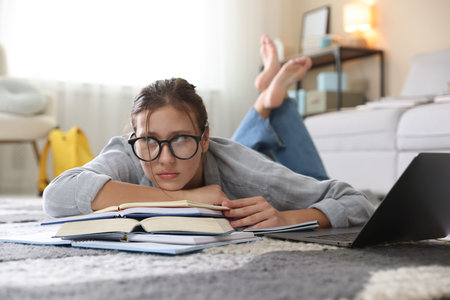 Worried student in glasses with laptop and books before exam on floor indoors