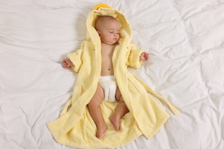Cute baby in bathrobe sleeping on bed, top view