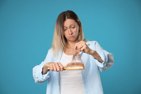 Alopecia problem. Sad woman taking her lost hair from brush on light blue backgroundの写真素材