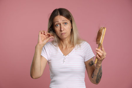 Alopecia problem. Sad woman holding brush with lost hair on pink backgroundの写真素材
