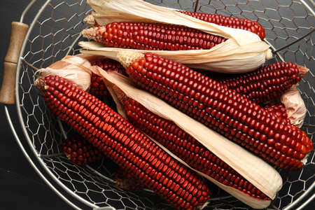 Ripe red corn cobs in metal basket on black wooden table, top view