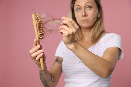 Alopecia problem. Worried woman taking her lost hair from brush on pink backgroundの写真素材