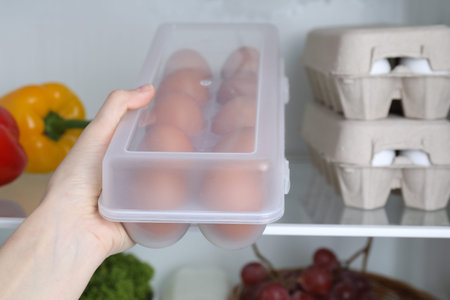 Woman putting container with raw chicken eggs into refrigerator, closeup