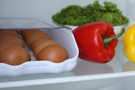 Container with raw chicken eggs and other food products on shelf in refrigerator, closeup