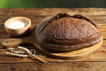 Loaf of fresh bread, flour and spikelets on wooden table against blurred background, closeup