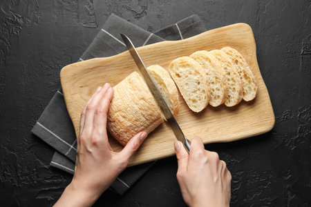 Woman cutting fresh crispy ciabatta at black table, top view