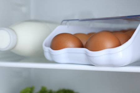 Container with raw chicken eggs and milk on shelf in refrigerator, closeup