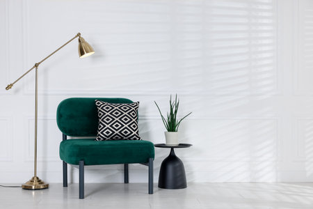 Stylish lamp, comfortable armchair with cushion and houseplant near white wall indoorsの写真素材