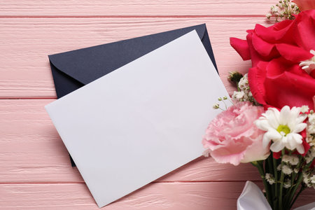 Blank card, envelope and flowers on pink wooden table, above view