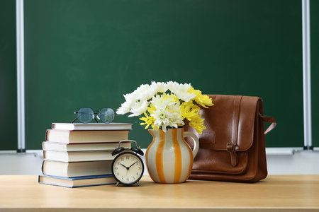 Happy Teacher's Day. Bag, books, alarm clock and vase with flowers on wooden table near chalkboardの写真素材