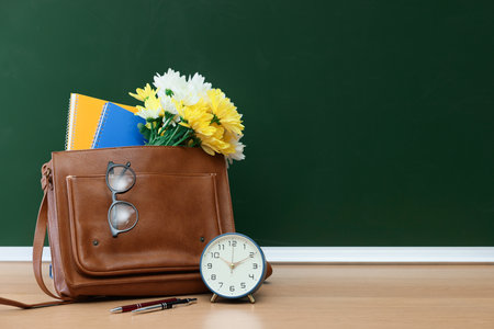 Happy Teacher's Day. Bag with stationery, alarm clock and flowers on wooden table near chalkboard, space for textの写真素材
