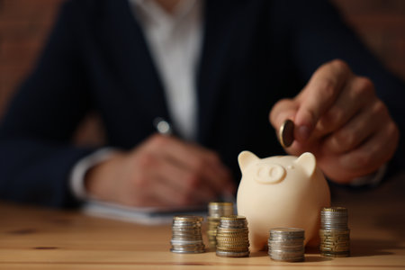 Man putting coin into piggy bank at wooden table indoors, selective focusの写真素材