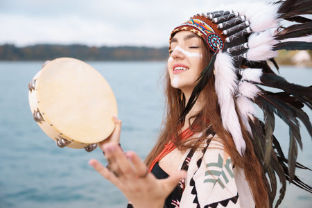 Smiling shaman woman in native headdress with face painting and tambourine performing ritual near river outdoorsの写真素材