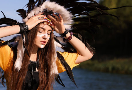Portrait of beautiful shaman woman in native American Indian headdress with face painting outdoors....