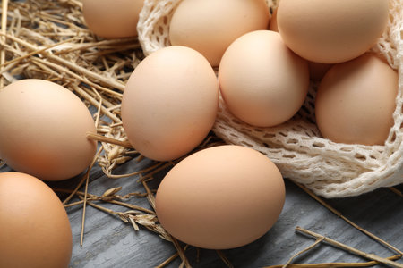 Raw chicken eggs, mesh bag and straw on gray wooden table, closeup