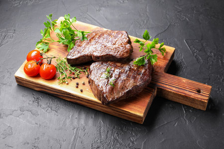 Delicious grilled beef steaks with herbs, peppercorns and tomatoes on dark textured table, closeupの写真素材