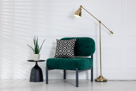 Stylish lamp, comfortable armchair with cushion and houseplant near white wall indoors. Interior designの写真素材