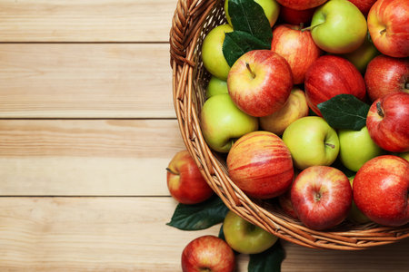 Fresh apples in wicker basket and green leaves on wooden table, top view. Space for textの写真素材