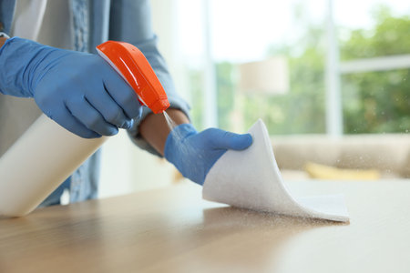 Woman spraying cleaning product while wiping wooden table with rag indoors, closeup