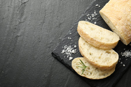 Cut crispy ciabatta, rosemary and salt on black textured table, top view. Space for text