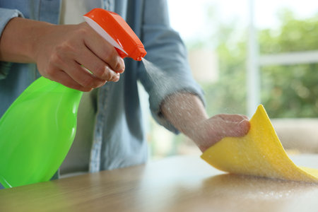 Woman spraying cleaning product while wiping wooden table with rag indoors, closeup