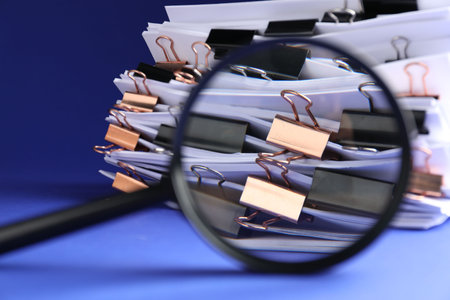 Magnifying glass and many documents with binder clips on blue background, closeupの写真素材