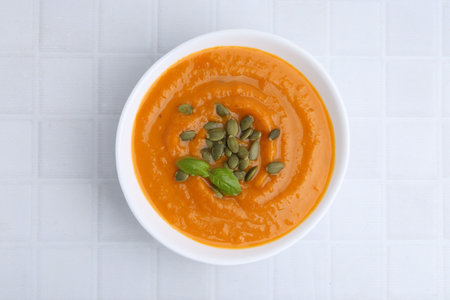Delicious pumpkin soup with seeds and basil in bowl on white tiled table, top viewの写真素材