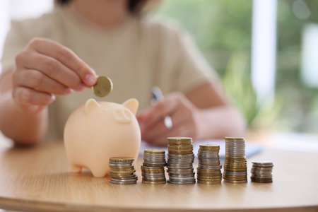 Woman putting coin into piggy bank at wooden table indoors, closeup
