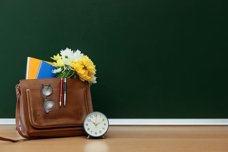 Happy Teacher's Day. Bag with stationery, alarm clock and flowers on wooden table near chalkboard, space for textの写真素材