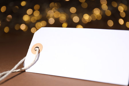 Blank tag on brown background with blurred lights, closeup. Bokeh effect