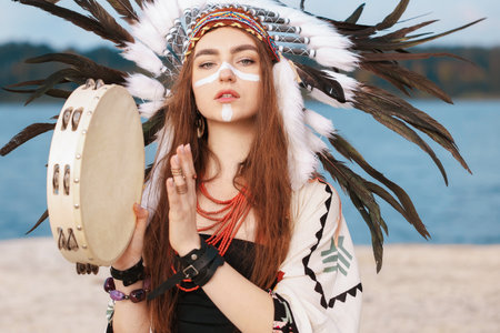 Beautiful shaman woman in native American Indian headdress with face painting, jewelry and tambourine performing ritual outdoorsの写真素材