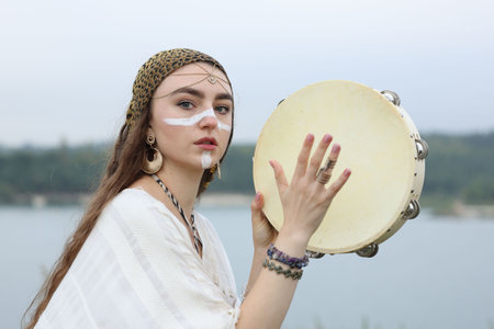 Beautiful shaman woman with face painting and tambourine performing ritual near river outdoorsの写真素材